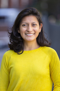 Diana, a Latinx woman with black, medium hair wearing a yellow sweater smiles in front of a blurred out street.